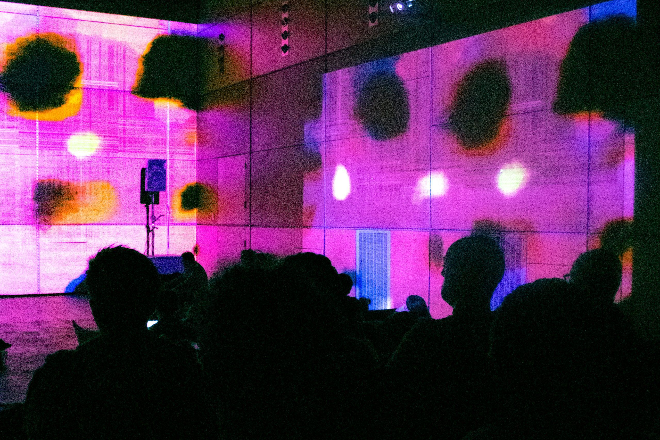 Audience silhouettes watch a vibrant, abstract projection with pink, purple, and orange hues and black patches on a large wall in a dimly lit room with speakers