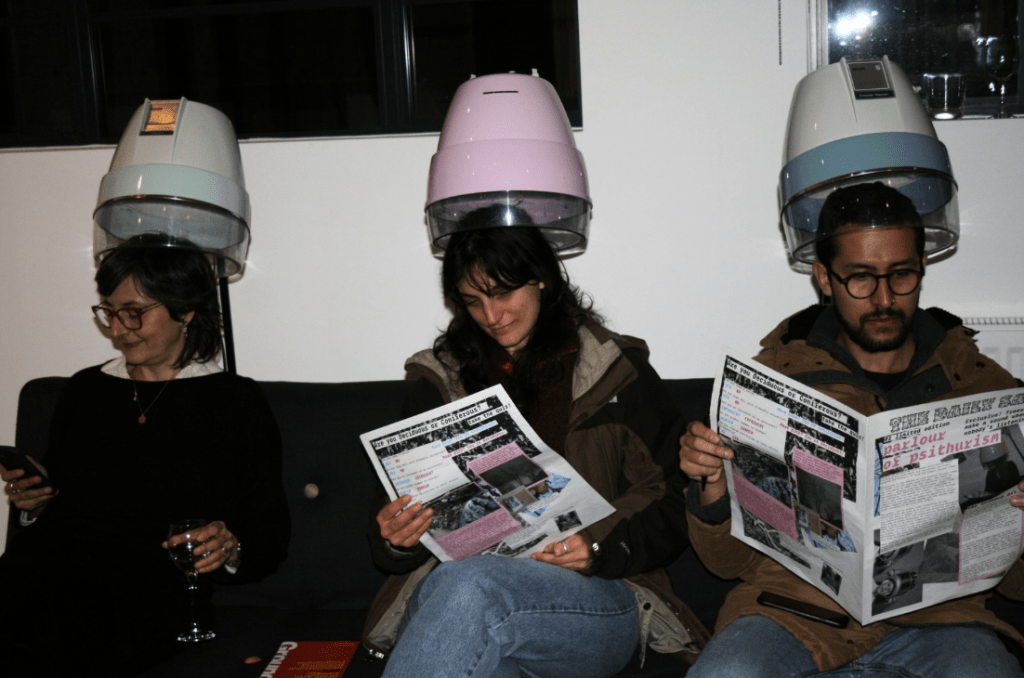 Three people sit on a sofa wearing vintage salon hairdryer hoods. Two are reading printed papers and one holds a phone and a glass of wine. The setting appears to be a relaxed gallery or event space with soft lighting and a casual atmosphere.