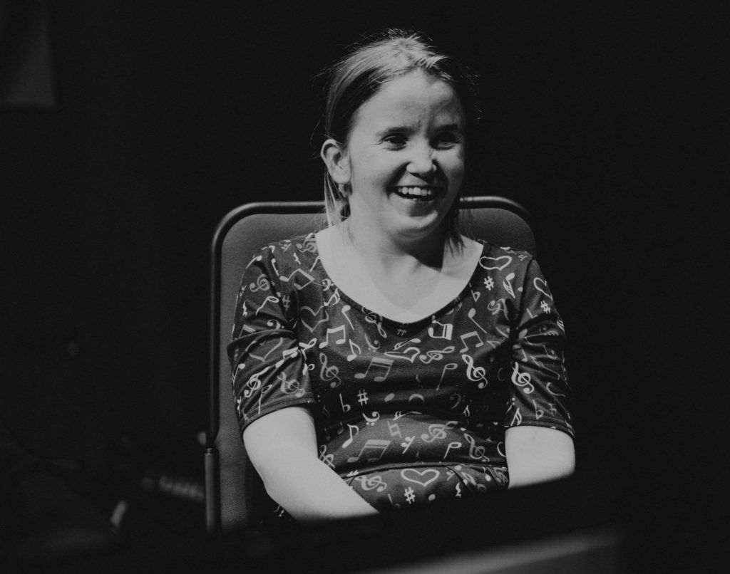 Sorcha sitting with two microphones on stands around her whilst she holds her phone into microphone during Beyond Borders Workshop at The Queen's Hall Edinburgh- she is wearing a dress with musical notes on it. The image is black and white.