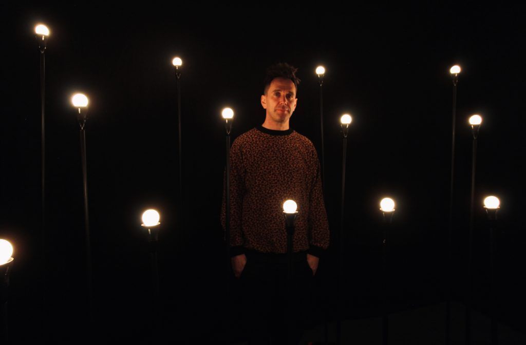 Many white light bulbs on stands, they are in lines and the background is black. The artist stand in the middle.