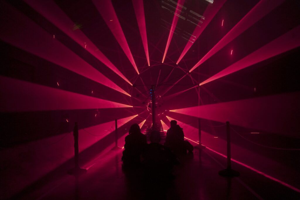 Three people sit on the floor in front of red lasers.