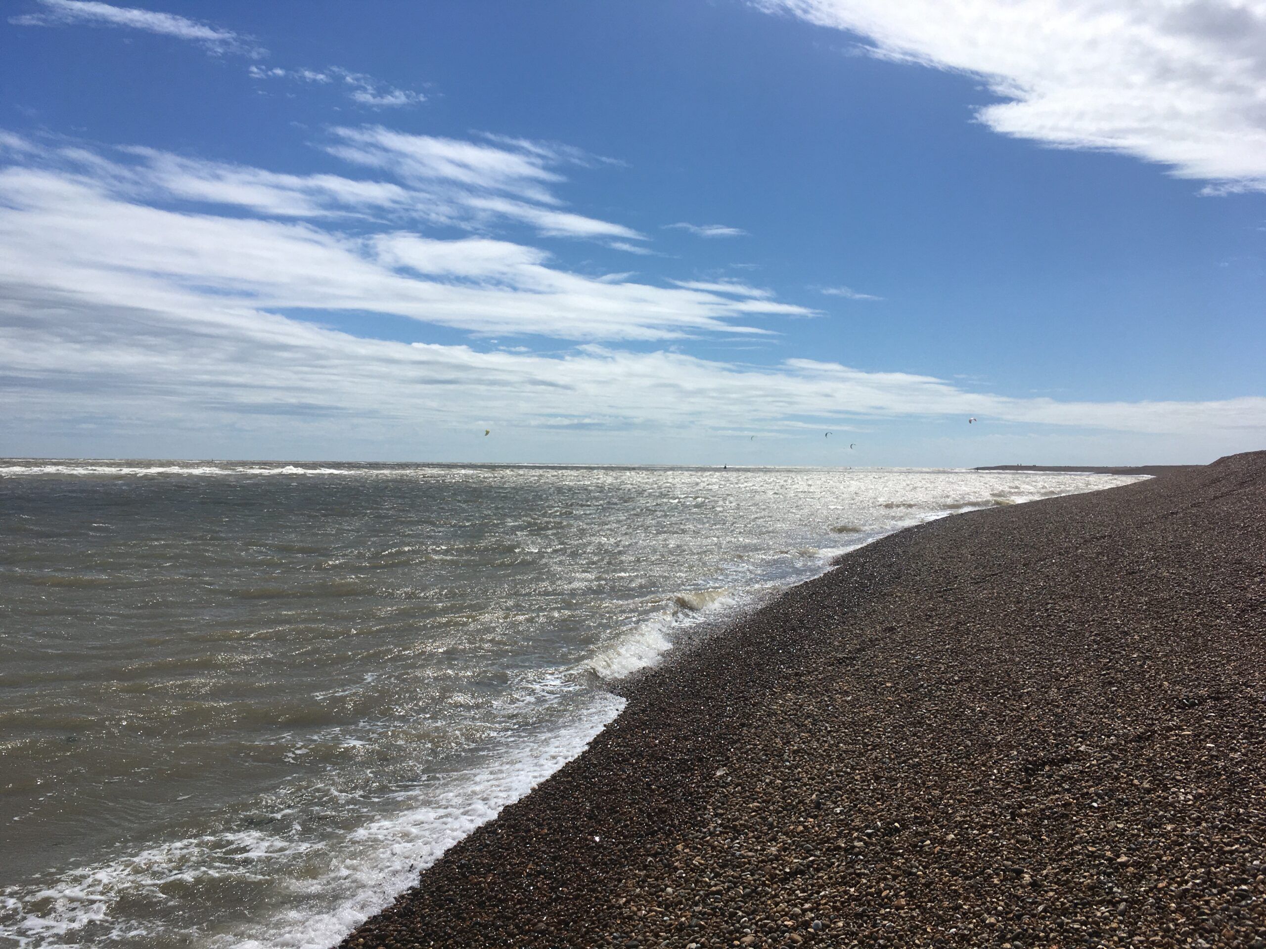 Shingle Street beach in Suffolk, a long shingle beach with blue skies and kite surfers in the grey sea.