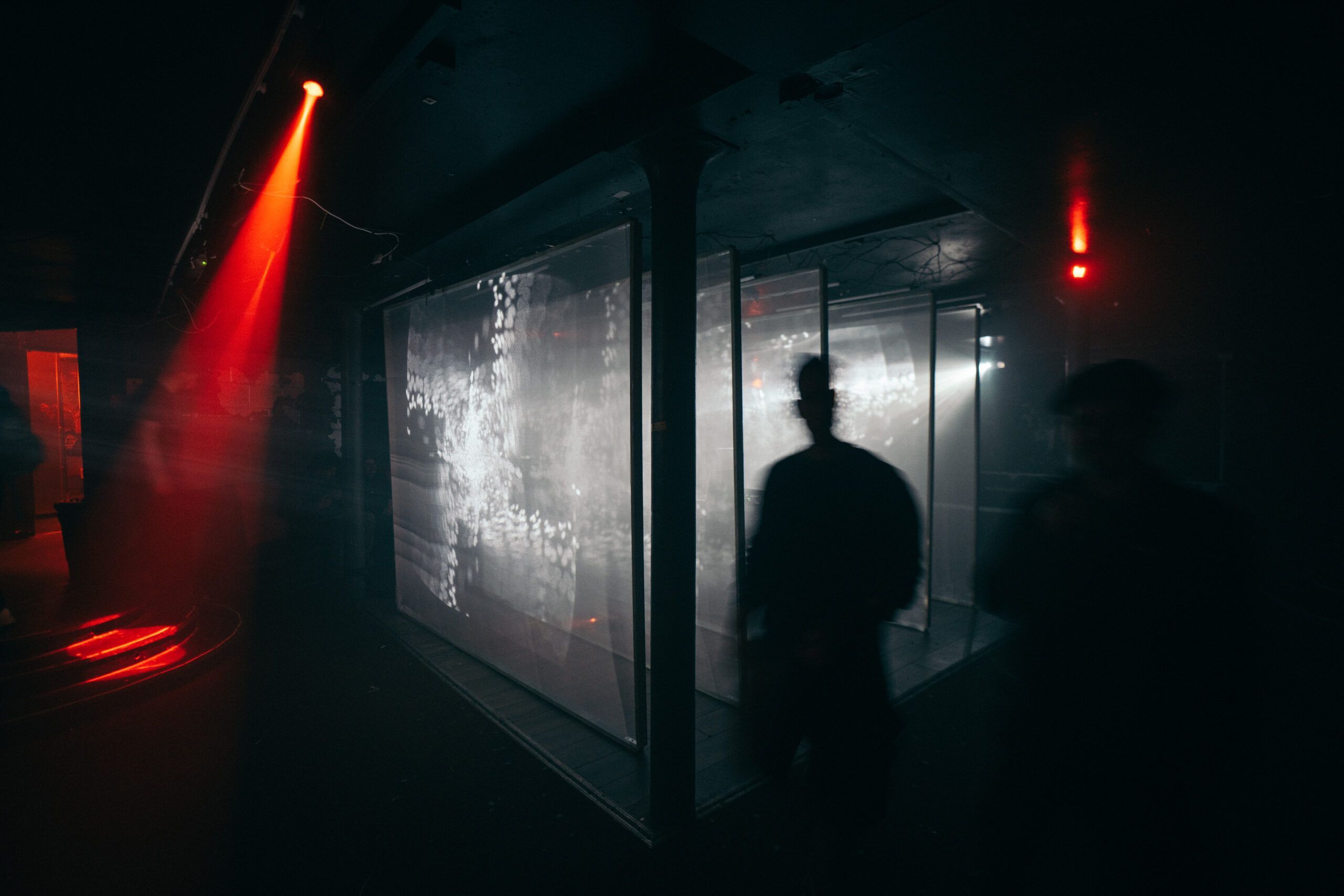 Audience members in silhouette view a large audiovisual installation in a darkened space, illuminated by red light and projected monochrome imagery.